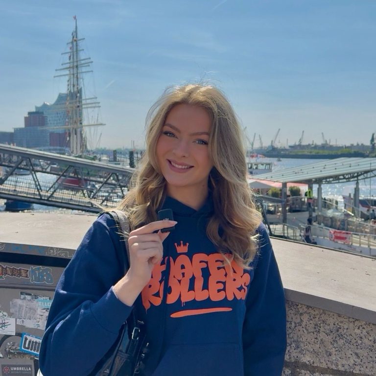 Farah Bär Junge Frau mit blonden Haaren in einem blauen Sweatshirt steht am Wasser.